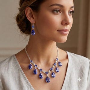 May include: A silver-toned necklace and matching drop earring set featuring multiple pear-shaped, deep blue faceted stones. The necklace consists of a delicate chain with dangling blue teardrop pendants, while each earring features a single matching blue teardrop stone suspended from a small circular stud.