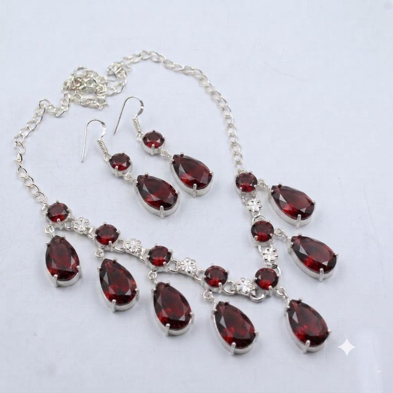 May include: A silver-toned necklace and matching pair of drop earrings featuring faceted, pear-shaped deep red stones. The necklace has a chain with multiple dangling red stone pendants, and the earrings have a single red stone drop design.