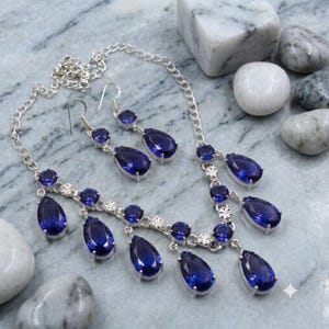 May include: A silver-toned necklace and matching drop earring set featuring faceted, pear-shaped deep blue stones and smaller round blue stones. The necklace has a delicate chain link design with multiple dangling blue stone pendants.
