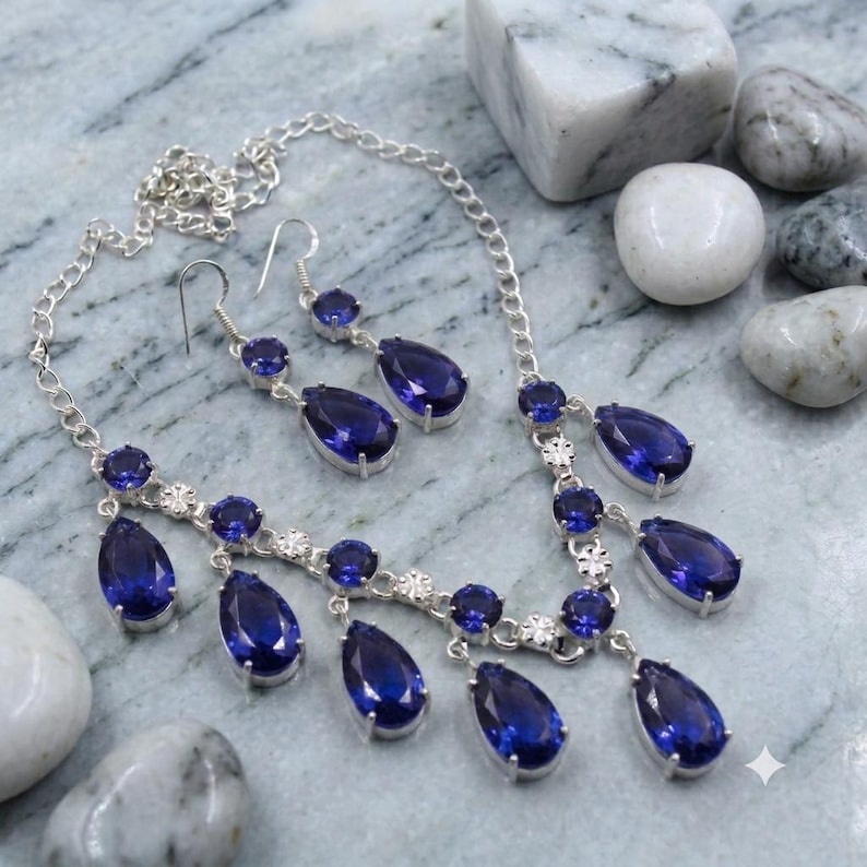 May include: A silver-toned necklace and matching drop earring set featuring faceted, pear-shaped deep blue stones and smaller round blue stones. The necklace has a delicate chain link design with multiple dangling blue stone pendants.