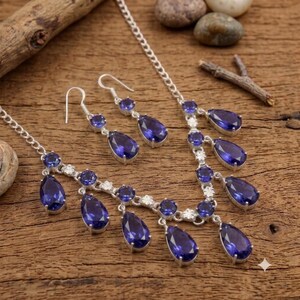 May include: A silver-toned necklace and matching pair of drop earrings featuring deep blue, pear-shaped gemstones. The necklace has a delicate chain with multiple dangling blue stones and small, clear, round accent stones set in silver metal.