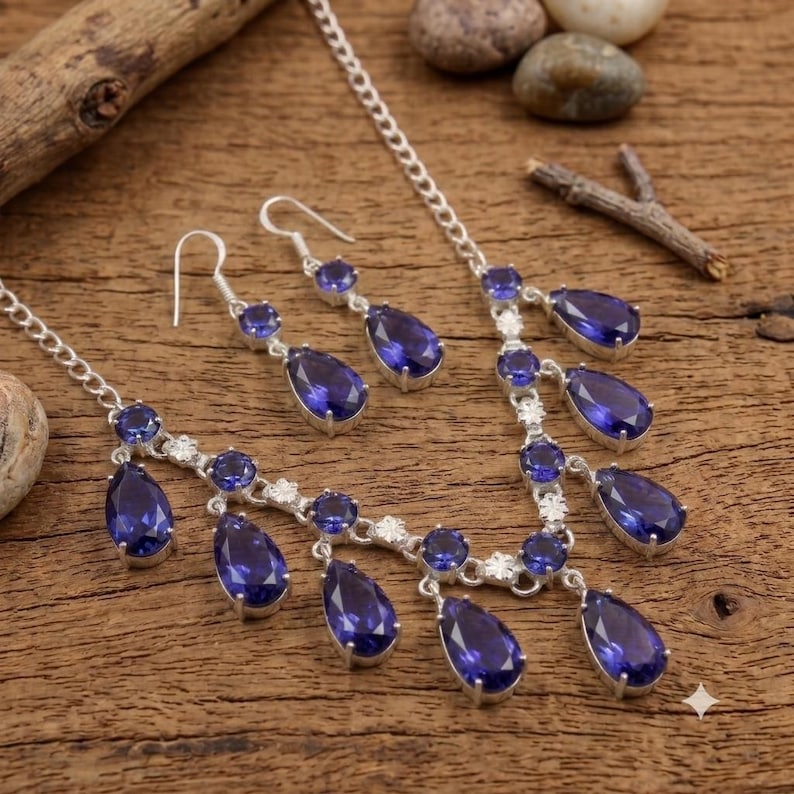 May include: A silver-toned necklace and matching pair of drop earrings featuring deep blue, pear-shaped gemstones. The necklace has a delicate chain with multiple dangling blue stones and small, clear, round accent stones set in silver metal.
