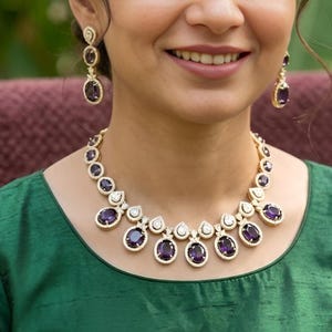 May include: A jewelry set consisting of a necklace and matching drop earrings. The necklace features a series of oval-shaped deep purple stones set in gold-toned metal, accented with small white stones. The earrings mirror this design.