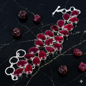 Puede incluir: Una pulsera de plata con múltiples piedras preciosas de granate rojo oscuro, talladas en forma de pera y dispuestas en doble fila, asegurada con un cierre de barra, acompañada de varias piedras de granate en bruto y oscuras.
