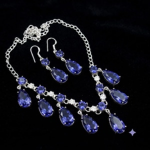 May include: A silver-toned necklace and matching pair of drop earrings featuring faceted, pear-shaped deep blue stones. The necklace has a delicate chain with multiple dangling blue stone pendants and small circular accents throughout the design.