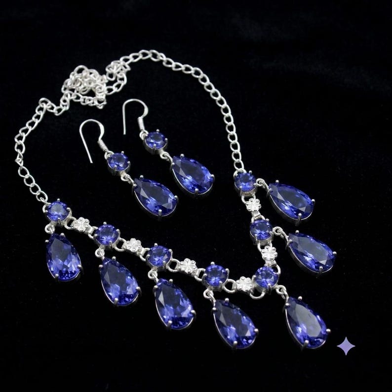 May include: A silver-toned necklace and matching pair of drop earrings featuring faceted, pear-shaped deep blue stones. The necklace has a delicate chain with multiple dangling blue stone pendants and small circular accents throughout the design.