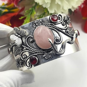 Puede incluir: Un brazalete ancho de plata con un cabujón central de cuarzo rosa ovalado, acentuado por una mariposa esculpida, una serpiente sinuosa y motivos florales. El diseño incluye varias piedras preciosas de granate rojo oscuro facetadas.