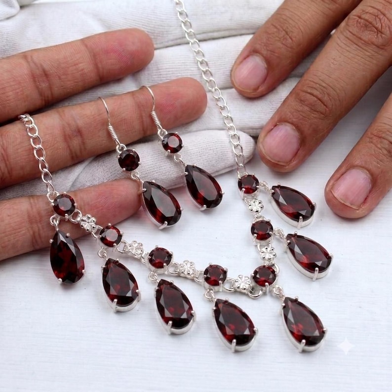 May include: A silver necklace and matching drop earrings set featuring faceted, deep red, pear-shaped garnet gemstones. The necklace consists of a delicate silver chain with multiple dangling garnet pendants, while each earring features a single garnet.
