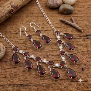 May include: A silver-toned necklace and matching pair of drop earrings featuring faceted, deep red, pear-shaped stones. The necklace has a delicate chain with multiple dangling stone pendants, and the earrings have a hook-style closure.