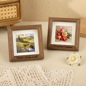 May include: Two square, wooden picture frames with landscape and floral art prints. One frame reads "A piece of my world" and the other reads "A moment I painted". A white flower and a decorative wooden box are also visible.
