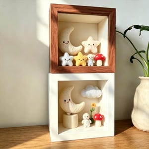 May include: Two shadow boxes, one with a dark wood frame and the other with a white frame, each containing felted celestial and woodland creatures. The top box features a smiling moon, stars, a mushroom, and a small figure. The bottom box has a moon, cloud, flower, mushroom, and figure.