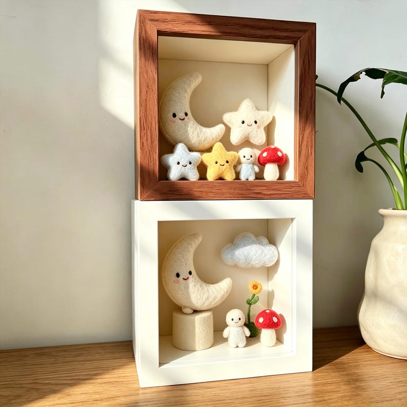 May include: Two shadow boxes, one with a dark wood frame and the other with a white frame, each containing felted celestial and woodland creatures. The top box features a smiling moon, stars, a mushroom, and a small figure. The bottom box has a moon, cloud, flower, mushroom, and figure.