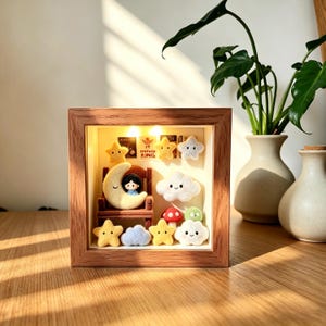 May include: A wooden shadow box with a whimsical night-themed scene. It features a crescent moon with a seated figure, stars, clouds, and a mushroom. The text "IT SHIVER KING" is visible. The scene is illuminated by small lights.