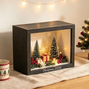 May include: A rectangular shadow box with a dark wood frame featuring a miniature Christmas scene inside. The display includes two green bottle brush trees, two small gift boxes with ribbons, red berries, and white snowflakes with warm lights.