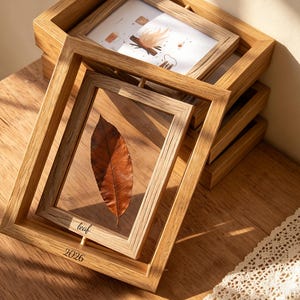 Może przedstawiać: Drewniane ramki na zdjęcia ułożone na drewnianej powierzchni. Jedna ramka przedstawia suszony liść, druga rysunek ogniska. Tekst "leaf" i "2026" jest widoczny na przedniej ramce. Ramki mają naturalne wykończenie słojów drewna.