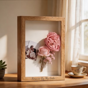 May include: A rectangular wooden shadow box frame with a light oak finish. Inside, a white backing displays two small photographs of a couple and a bouquet of preserved pink roses and baby's breath. The text Forever By My Side is engraved.