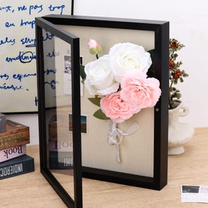May include: A black rectangular shadow box frame with a glass front door, featuring a beige linen-textured interior backing. Inside, a bouquet of artificial white and light pink roses is attached, with a small white ribbon tied at the stems.