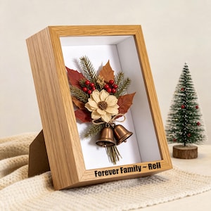 May include: A light wood shadow box frame containing a dried floral arrangement with red berries, green pine needles, and two bronze-colored bells. The text "Forever Family ~ Rell" is printed in black on the bottom edge of the wooden frame.
