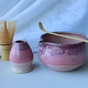 May include: A matcha tea set comprising a bamboo whisk, a small pink ceramic tea bowl, a matching tea cup, and a bamboo scoop. The tea bowl and cup have a two-tone pink glaze. The whisk is made of light-coloured bamboo.
