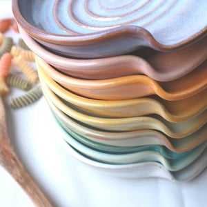Op de afbeelding: Een stapel keramische lepelhouders in diverse pastelkleuren, waaronder lichtblauw, roze, oranje, geel, groen en wit. Elke lepelhouder heeft een ronde kom met een inkeping voor de steel en is afgewerkt met een gladde, geglazuurde laag.