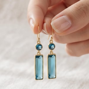 May include: A pair of gold-tone drop earrings featuring a small round blue gemstone set above a larger rectangular blue gemstone. The earrings hang from thin gold-tone hooks and display a clear, faceted design with a polished metallic finish.