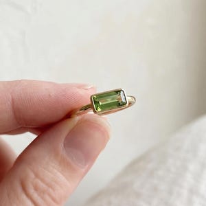 Könnte beinhalten: Ein Goldring mit einem rechteckigen, im Baguetteschliff geschliffenen grünen Edelstein in einer Zargenfassung. Der Ring verfügt über eine glatte, polierte Goldschiene, die den Finger umschließt und die lebendige, olivgrüne Farbe betont.