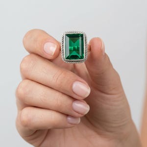 Peut inclure: Bague en argent ornée d'une grande pierre verte de taille émeraude. La bague présente une bordure texturée en forme de corde autour de la pierre. Fond blanc.