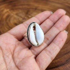 Könnte beinhalten: Ein Silberring mit einer großen, oval geformten weißen Muschel. Die Muschel ist in einem Silberband mit einem Perlenrand gefasst. Der Ring wird auf einer Hand präsentiert.