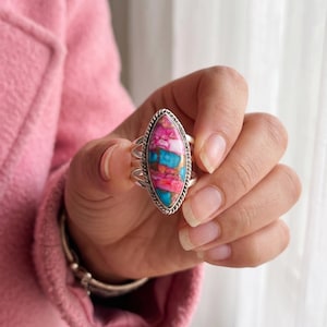 Könnte beinhalten: Ein marquiseförmiger Ring mit einer Mosaikeinlage aus rosa und türkisfarbenen Steinen, gefasst in ein Silberband mit gedrehter Kordelbordüre. Der Stein zeigt eine lebendige Mischung aus hellrosa und blauen Segmenten, getrennt durch dunkle Linien.