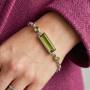 Può includere: Un braccialetto d'argento con una grande gemma rettangolare verde chiaro e pietre verdi rotonde più piccole. Il braccialetto è su un polso, con un tessuto rosa-viola sullo sfondo.
