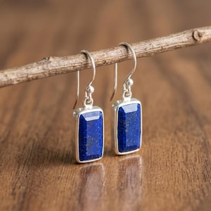 May include: A pair of silver drop earrings with rectangular, deep blue lapis lazuli stones. The earrings are suspended from simple hooks and are displayed on a wooden branch against a blurred wooden background.