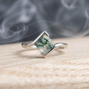 Könnte beinhalten: Ein Silberring mit einem drachenförmigen Moosachat-Edelstein. Der Stein ist durchscheinend und weist deutliche grüne, farnartige Einschlüsse auf. Der Metallring hat eine glatte, polierte Oberfläche und ist geschwungen gefasst.