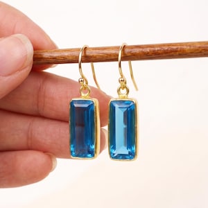 May include: A pair of gold-colored earrings with rectangular, bright blue gemstones. The gemstones are set in a gold bezel and have a polished finish. The earrings are held up by a wooden stick.