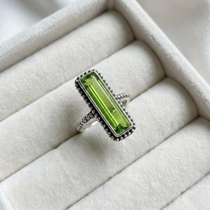Könnte beinhalten: Ein Silberring mit einem rechteckigen, hellgrünen Edelstein. Der Edelstein ist in einem Silberband mit einem Perlenrand gefasst. Der Ring wird auf einer weißen, strukturierten Oberfläche präsentiert.