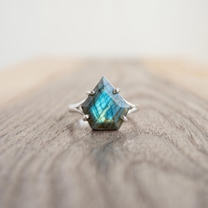 May include: A silver ring featuring a pentagonal labradorite gemstone with a faceted surface. The stone displays iridescent blue and gold flashes within its structure. The metal band is thin and polished, holding the stone in a prong setting.