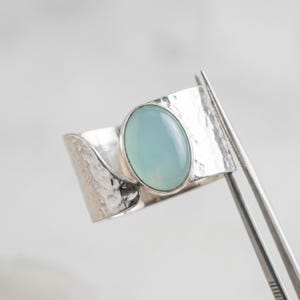 Könnte beinhalten: Ein breiter Silberring mit gehämmertem Muster und einem großen, ovalen, hellblauen Edelstein. Der Ring wird von einer Pinzette gehalten, die das detaillierte Design und die Handwerkskunst des Schmucks zeigt.