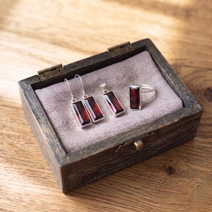Könnte beinhalten: Ein Schmuckset bestehend aus zwei rechteckigen Ohrhängern, einem passenden rechteckigen Anhänger und einem Ring, alle mit tiefroten facettierten Steinen in silberfarbenem Metall gefasst, präsentiert in einer kleinen dunklen Holzschatulle mit Stoff.