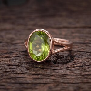 Könnte beinhalten: Ein Ring aus Roségold mit einem großen, oval geschliffenen, hellgrünen Peridot-Edelstein in einer glatten Fassung. Die Ringschiene hat ein geteiltes Design, das sich nach hinten verjüngt, und besticht durch eine polierte metallische Oberfläche.