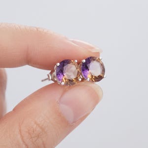 Könnte beinhalten: Ein Paar runde Ohrstecker mit facettierten Ametrin-Edelsteinen, die einen Farbverlauf von Violett bis Goldbraun aufweisen. Die Steine sind in vierzinkige Fassungen aus silberfarbenem Metall eingefasst und besitzen einen klaren Schliff.