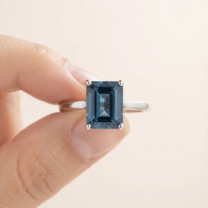 May include: A silver-toned ring featuring a large, rectangular emerald-cut dark blue gemstone set in a four-prong setting. The band is smooth and polished, showcasing a clean, minimalist design that highlights the deep blue color of the stone.