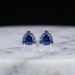 Könnte beinhalten: Ein Paar Ohrstecker mit dreieckig geschliffenen, tiefblauen Edelsteinen, die in silberfarbenen Metallfassungen sitzen. Jeder Ohrstecker hat drei einzelne Krappen, die den facettierten blauen Stein sicher halten und eine geometrische Form bilden.