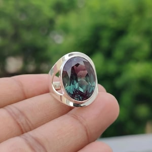 Pode incluir: Um anel de prata com uma grande pedra preciosa oval que muda de cor, exibindo tons de roxo e verde-azulado. O anel tem uma banda larga com um design recortado em cada lado, oferecendo um estilo moderno e elegante.