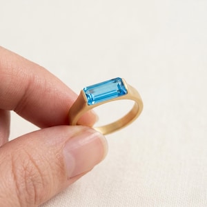Könnte beinhalten: Ein goldener Ring mit einem rechteckigen, hellblauen Edelstein. Der Ring hat ein schlichtes, modernes Design mit glatter, matter Oberfläche. Der Edelstein ist in einer Lünettenfassung gefasst, und das Band hat eine goldene Farbe.