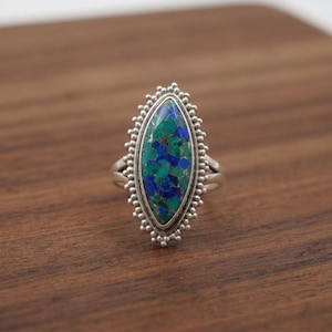 Könnte beinhalten: Ein silberner Ring mit einem großen, oval geformten Stein. Der Stein weist eine Mischung aus blauen und grünen Farbtönen mit einem gesprenkelten Muster auf. Der Ring hat einen detaillierten, perlenbesetzten Rand und ein geteiltes Schaftdesign. Der Ring liegt auf einer Holzoberfläche.