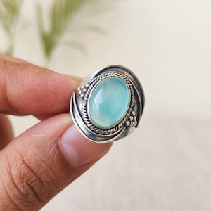 Könnte beinhalten: Ein Silberring mit einem zentralen, ovalen, glatten, hellblauen Cabochon-Stein. Die Fassung umfasst einen gedrehten Kordelrand und kleine dekorative Metallperlen an den Seiten, mit einem polierten, oxidierten Silberband-Finish.
