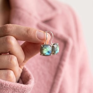 Puede incluir: Un par de pendientes colgantes de plata con piedras de labradorita de corte cuadrado que muestran un brillo iridiscente azul y verde. Las piedras están sujetas por cuatro garras y unidas a ganchos de plata curvados para el oído.