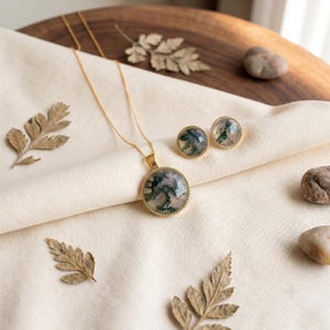 Elegant Moss Agate jewelry set displayed inside a luxury wooden jewelry box with soft pink velvet lining. The set includes a matching round Moss Agate pendant necklace and coordinating stud earrings, each featuring natural green moss-like inclusions within translucent white gemstones. The stones are bezel-set in gold-plated frames, creating a timeless and sophisticated look. This nature-inspired handmade jewelry set is perfect for weddings, bridesmaids, special occasions, and everyday wear,