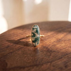 Può includere: Un anello in oro con una pietra preziosa di agata muschiata ovale allungata con inclusioni dendritiche verdi. La fascia presenta una delicata trama a perline e la pietra è incastonata in una lunetta d'oro liscia, creando un gioiello raffinato.