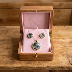 Elegant Moss Agate jewelry set displayed inside a luxury wooden jewelry box with soft pink velvet lining. The set includes a matching round Moss Agate pendant necklace and coordinating stud earrings, each featuring natural green moss-like inclusions within translucent white gemstones. The stones are bezel-set in gold-plated frames, creating a timeless and sophisticated look. This nature-inspired handmade jewelry set is perfect for weddings, bridesmaids, special occasions, and everyday wear,