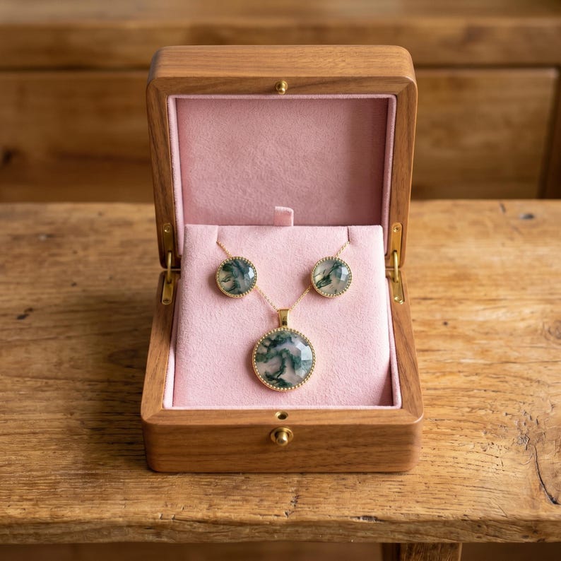 Elegant Moss Agate jewelry set displayed inside a luxury wooden jewelry box with soft pink velvet lining. The set includes a matching round Moss Agate pendant necklace and coordinating stud earrings, each featuring natural green moss-like inclusions within translucent white gemstones. The stones are bezel-set in gold-plated frames, creating a timeless and sophisticated look. This nature-inspired handmade jewelry set is perfect for weddings, bridesmaids, special occasions, and everyday wear,